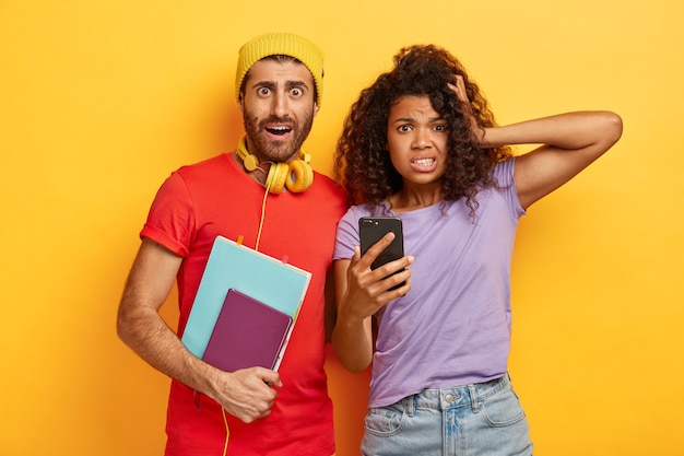 Un joven y un hombre desconcertados y conmocionados usan el teléfono celular, tienen fecha límite para preparar el proyecto, estudian juntos, usan camisetas casuales brillantes, miran con expresión omg