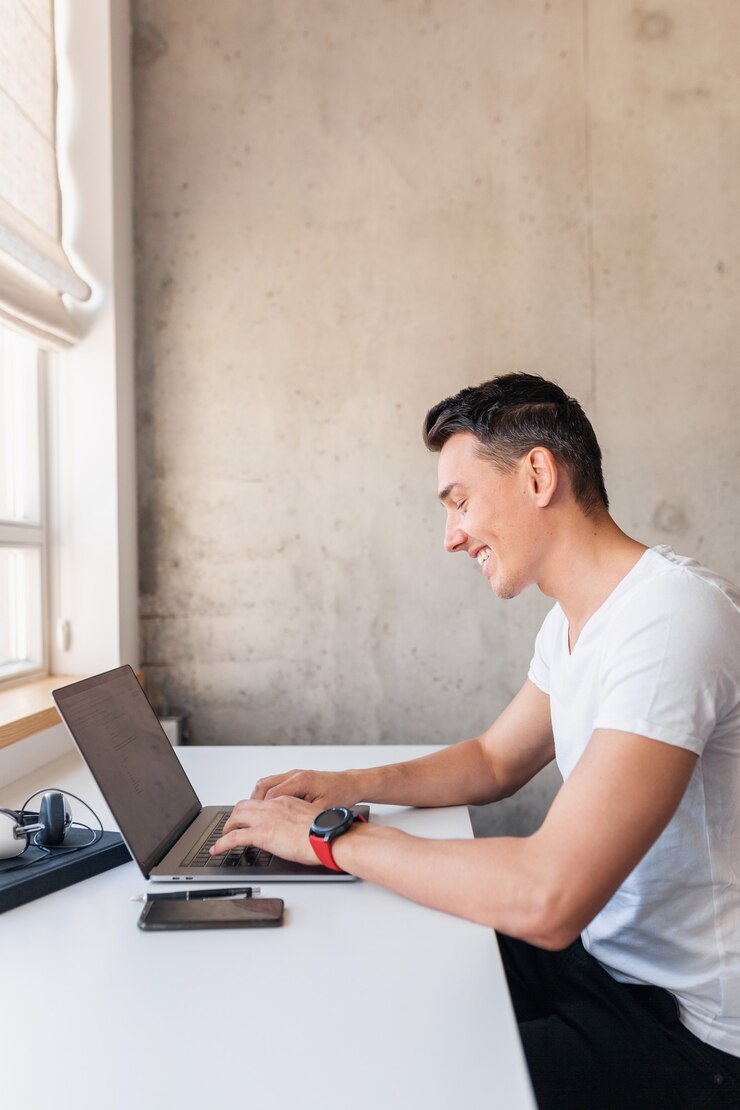 Foto gratuito joven hombre guapo y sonriente con ropa casual sentado en la mesa trabajando en una computadora portátil