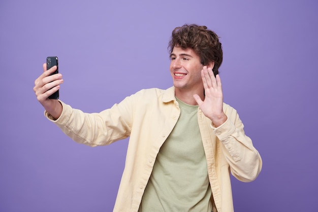 Foto gratuita joven hombre guapo sonriente saludando y saludando con la mano mientras hace una videollamada en un teléfono inteligente