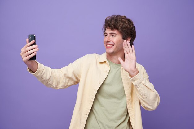 Joven hombre guapo sonriente saludando y saludando con la mano mientras hace una videollamada en un teléfono inteligente