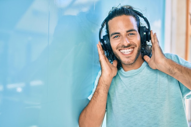 Joven hombre hispano sonriendo feliz escuchando música usando auriculares en la ciudad