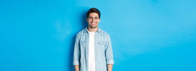 Joven hombre moderno con gafas y ropa informal de pie contra un fondo azul sonriendo feliz cuando llegó