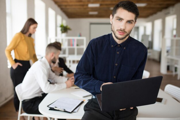 Joven hombre de negocios con barba en camisa mirando cuidadosamente en cámara sosteniendo una computadora portátil en las manos mientras pasa tiempo en la oficina con colegas en el fondo