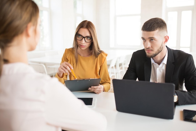 Joven hombre de negocios con computadora portátil y mujer de negocios en anteojos mirando cuidadosamente al solicitante Jóvenes empleadores gastando una entrevista de trabajo en una oficina moderna