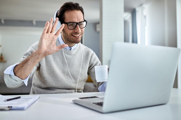 Joven hombre de negocios feliz saludando mientras tiene una reunión en línea sobre una computadora portátil en casa