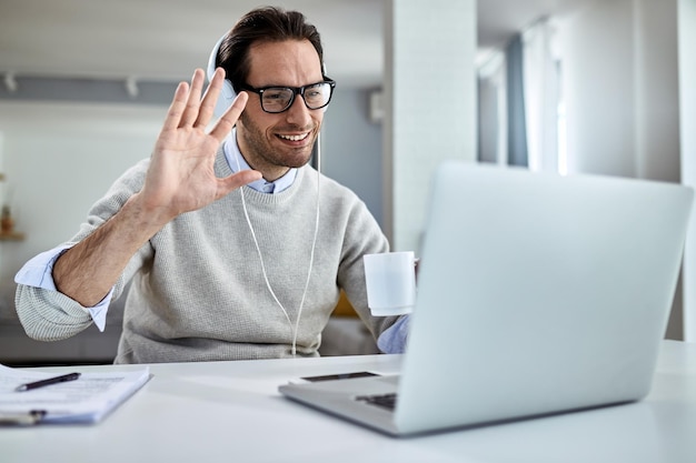 Joven hombre de negocios feliz saludando mientras tiene una reunión en línea sobre una computadora portátil en casa