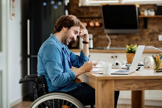 Joven hombre de negocios feliz en silla de ruedas trabajando en casa y enviando mensajes de texto por teléfono móvil.