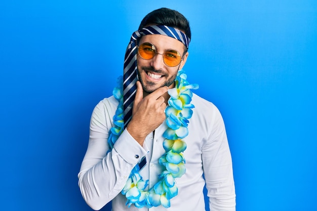 Foto gratuita joven hombre de negocios hispano con estilo divertido de fiesta con corbata en la cabeza mirando con confianza a la cámara sonriendo con los brazos cruzados y la mano levantada en la barbilla pensando en positivo