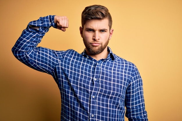 Joven hombre de negocios rubio con barba y ojos azules con camisa sobre fondo amarillo Persona fuerte que muestra músculo del brazo seguro y orgulloso del poder