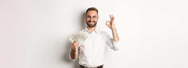 Un joven hombre de negocios satisfecho que muestra dinero hace bien el signo de ganar dinero de pie sobre fondo blanco