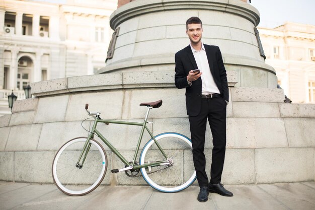 Joven hombre de negocios sonriente con traje negro clásico y camisa blanca con auriculares inalámbricos sosteniendo el celular en la mano mientras mira soñadoramente a la cámara con una bicicleta retro cerca de la calle