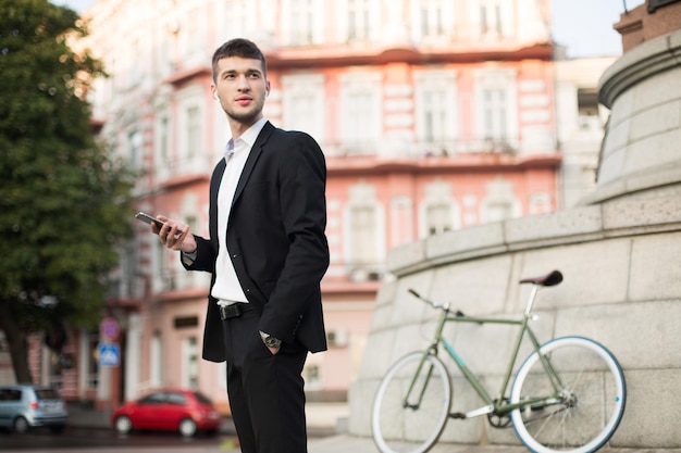 Joven hombre de negocios con traje negro clásico y camisa blanca con auriculares inalámbricos sosteniendo el celular en la mano mientras mira cuidadosamente a un lado con una bicicleta retro en el fondo al aire libre