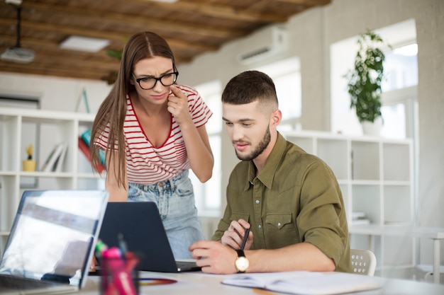 Joven hombre pensativo con camisa y mujer con camiseta a rayas y anteojos trabajando soñadoramente junto con una computadora portátil Gente de negocios creativa que pasa tiempo en el trabajo en una oficina moderna y acogedora