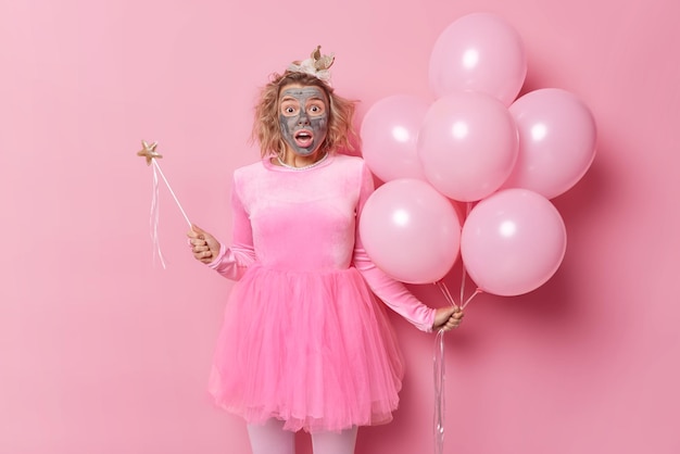 Una joven impresionada y conmocionada aplica una máscara de belleza reacciona ante algo increíble usa una diadema de vestido festivo sostiene un montón de globos de helio inflados y una varita mágica entretiene a los niños en la fiesta