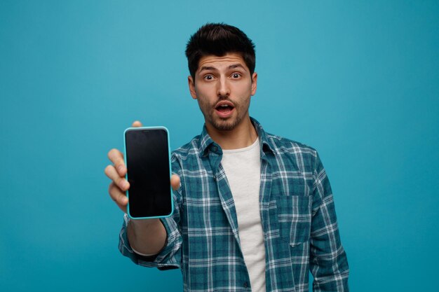 Joven impresionado mirando a la cámara estirando el teléfono móvil hacia la cámara aislada en el fondo azul