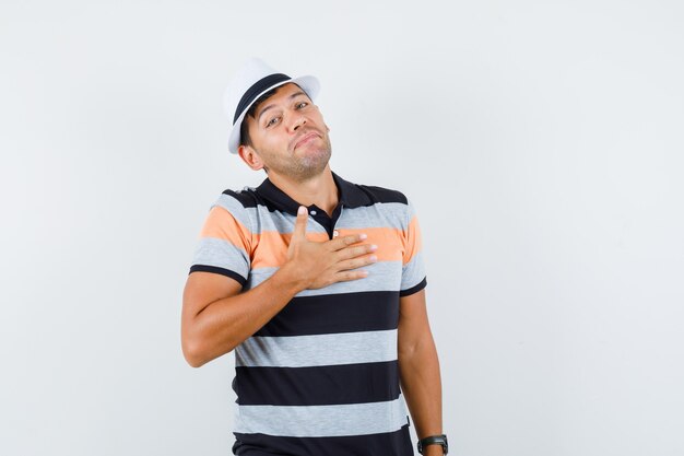 Joven inclinando la cabeza con la mano en el pecho en camiseta y sombrero y luciendo lindo