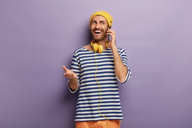 Joven inconformista habla casualmente con un amigo a través de un teléfono inteligente, habla de algo divertido que sucedió con él, tiene una expresión de cara feliz, usa un atuendo elegante, escucha música en auriculares. Comunicación