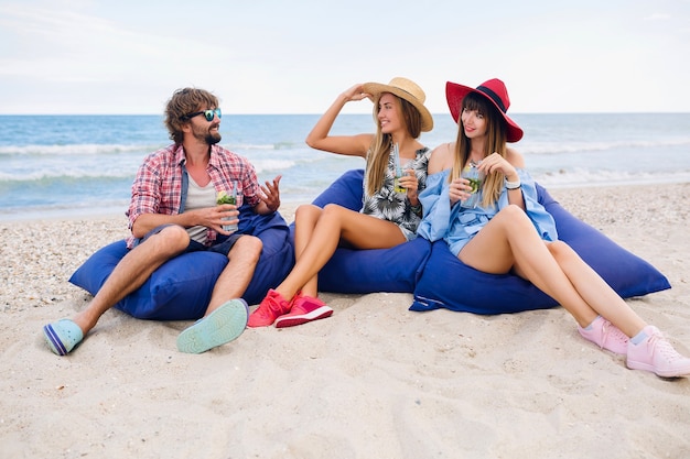 Joven inconformista sonriendo compañía de amigos felices de vacaciones sentados en bolsas de frijoles en una fiesta en la playa, bebiendo un cóctel mojito