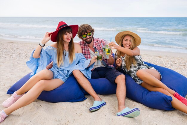 Joven inconformista sonriente compañía de amigos felices en vacaciones de verano sentado en la playa en bolsas de frijoles, brindando, riendo, bebiendo cócteles mojito