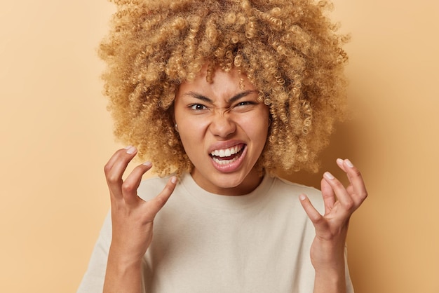 Una joven indignada con gestos de pelo rizado y tupido frunce el ceño frunciendo el ceño expresa emociones negativas vestida con una camiseta informal aislada sobre un fondo beige hace muecas por algo ridículo