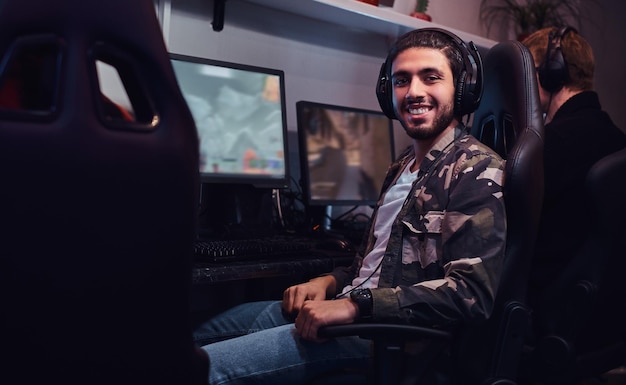 Un joven indio alegre con una camisa militar sentado en una silla de jugador y mirando una cámara en un club de juegos o en un cibercafé.