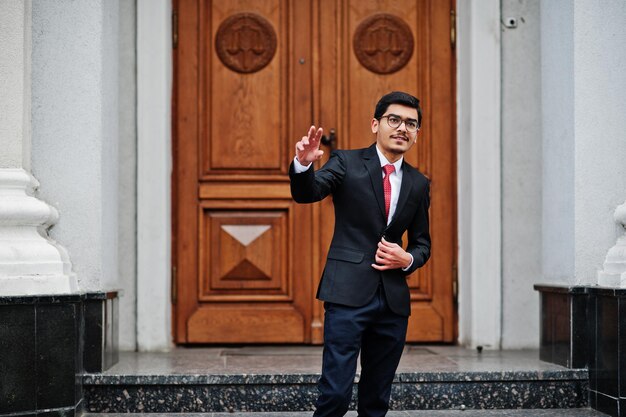 Joven indio con anteojos vestido con traje negro y corbata roja posado al aire libre contra la puerta del edificio muestra dos dedos