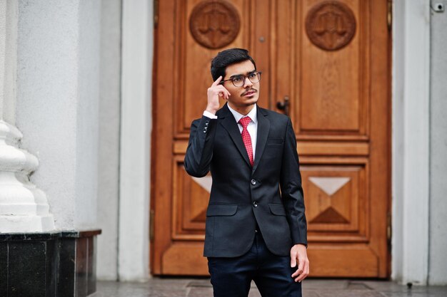 Un joven indio con anteojos vestido con traje negro y corbata roja posado al aire libre contra la puerta del edificio muestra que su pensamiento