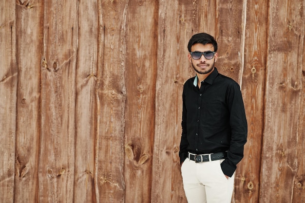 Un joven indio casual con camisa negra y gafas de sol posó contra un fondo de madera
