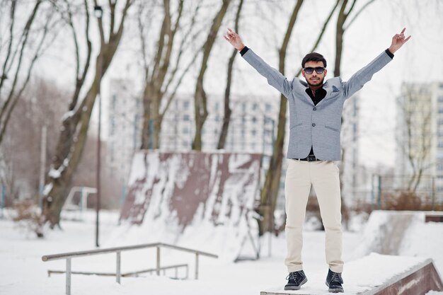 Joven indio casual con chaqueta plateada y gafas de sol mostrando el dedo de la roca con las manos levantadas y posando en el día de invierno