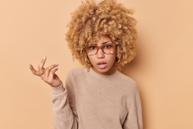 Una joven infeliz de cabello rizado se siente indignada, mantiene la mano levantada, se siente despistada, vestida con un suéter informal aislado sobre un estudio beige, no puede entender algo. Entonces, ¿qué dijiste?