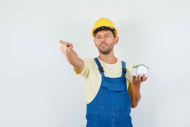 Joven ingeniero apuntando hacia afuera y sosteniendo el modelo de la casa en vista frontal uniforme.