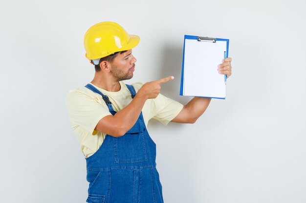 Joven ingeniero apuntando al portapapeles en uniforme y mirando enfocado. vista frontal.