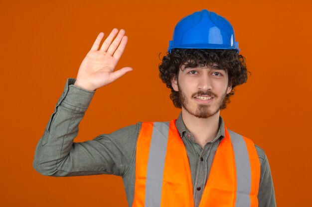 Joven ingeniero barbudo guapo con casco de seguridad y chaleco haciendo gesto de saludo saludando con la mano sonriendo sobre la pared naranja aislada
