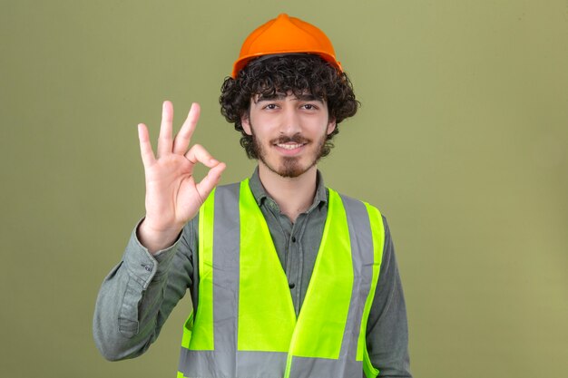 Joven ingeniero barbudo guapo con casco de seguridad y chaleco sonriendo haciendo aceptar firmar de pie sobre la pared verde aislada