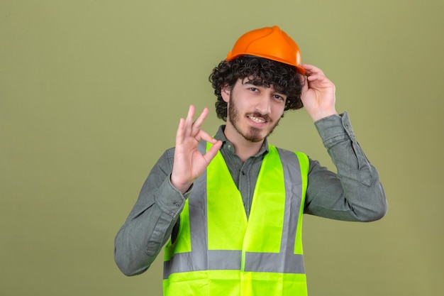 Joven ingeniero barbudo guapo con casco de seguridad y chaleco sonriendo haciendo aceptar firmar de pie sobre la pared verde aislada