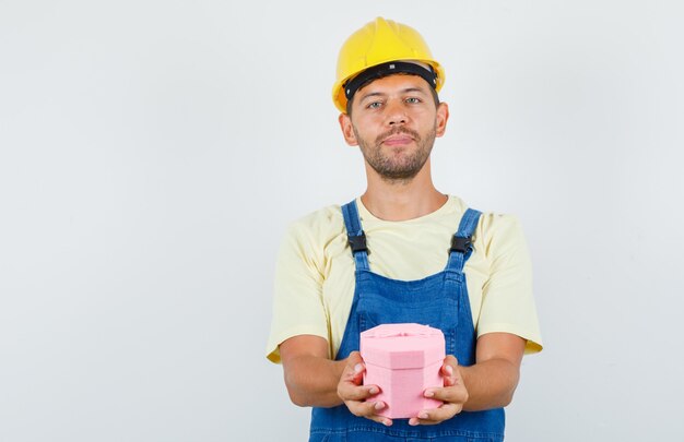 Joven ingeniero con caja de regalo en uniforme y alegre. vista frontal.