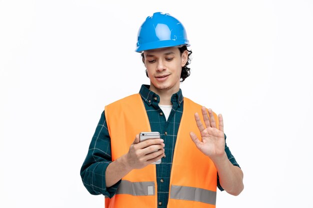 Joven ingeniero complacido con casco de seguridad y chaleco de seguridad hablando por videollamada haciendo un gesto de hola aislado en fondo blanco