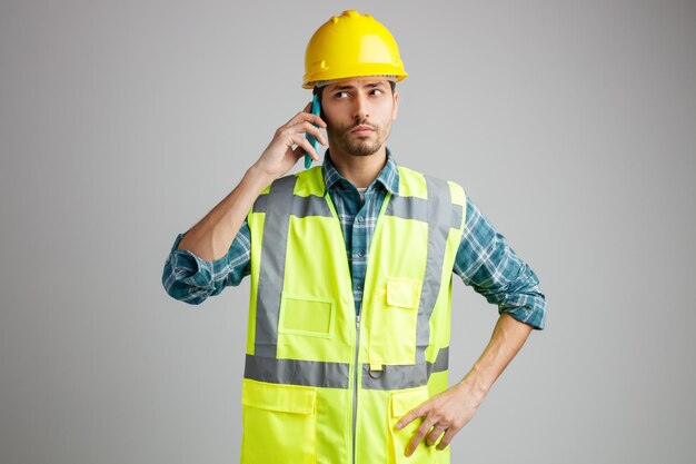 Joven ingeniero confiado usando casco de seguridad y uniforme manteniendo la mano en la cintura mirando al lado mientras habla por teléfono aislado en fondo blanco