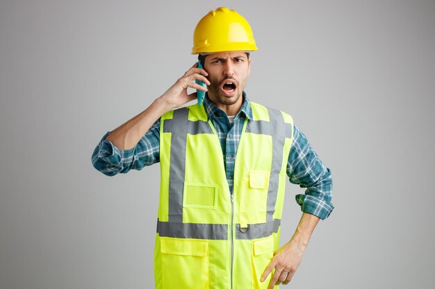 Joven ingeniero enojado con casco de seguridad y uniforme mirando a la cámara hablando por teléfono mientras mantiene la mano en la cintura aislada en el fondo blanco