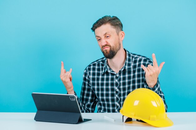 El joven ingeniero está mostrando gestos de rock con las manos sentado frente a la tableta sobre fondo azul.