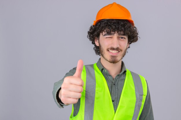 Joven ingeniero guapo con barba guiñando un ojo con una sonrisa en la cara mostrando el pulgar hacia arriba sobre la pared blanca aislada