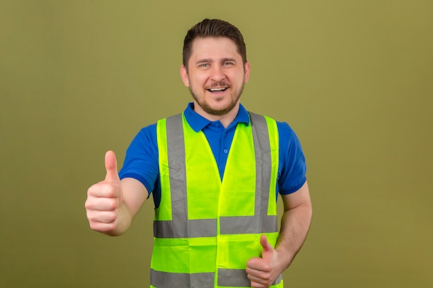 Foto gratuita joven ingeniero hombre vestido con chaleco de construcción mirando a cámara con gran sonrisa y cara feliz mostrando el pulgar hacia arriba sobre fondo verde aislado