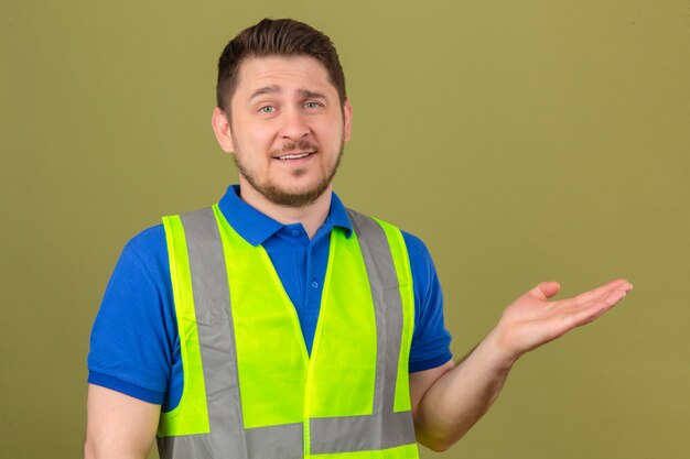 Joven ingeniero hombre vestido con chaleco de construcción mirando a la cámara presentando y apuntando con la palma de la mano sobre fondo verde aislado