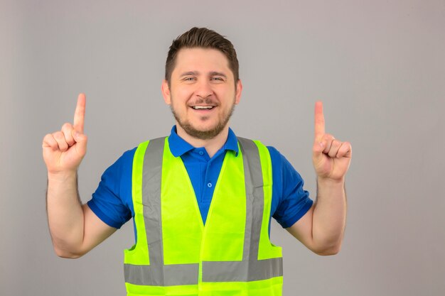 Joven ingeniero hombre vestido con chaleco de construcción mirando a la cámara sonriendo alegre apuntando hacia arriba con los dedos sobre fondo blanco aislado