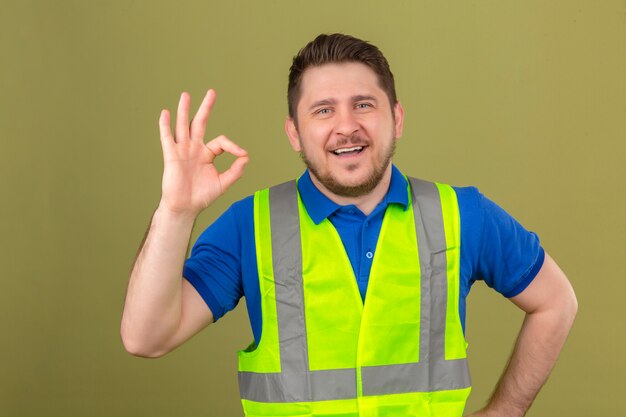 Joven ingeniero hombre vestido con chaleco de construcción mirando a la cámara sonriendo alegre haciendo bien firmar sobre fondo verde aislado