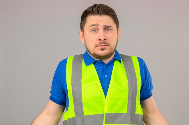 Joven ingeniero hombre vestido con chaleco de construcción que parece confundido sin tener idea de qué hacer parado sobre fondo blanco aislado