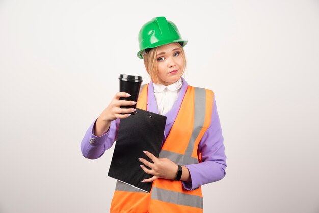 Joven ingeniero industrial en uniforme con portapapeles y taza negra sobre pared blanca.