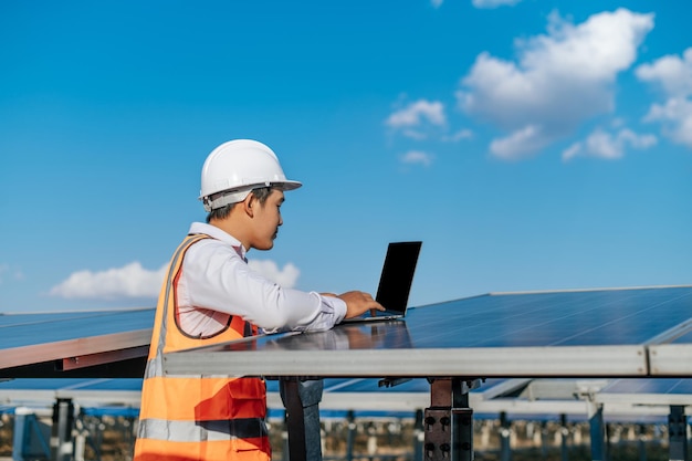 Un joven ingeniero inspector asiático usa una computadora portátil que trabaja en una granja solar Supervisor técnico masculino con casco blanco Comprobación del funcionamiento del panel solar y fotovoltaico en el espacio de copia de la estación