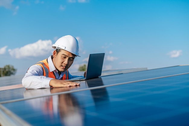 Un joven ingeniero inspector asiático usa una computadora portátil que trabaja en una granja solar Supervisor técnico masculino con casco blanco Comprobación del funcionamiento del panel solar y fotovoltaico en el espacio de copia de la estación