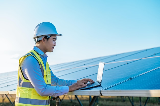 Un joven ingeniero inspector asiático usa una computadora portátil que trabaja en una granja solar Supervisor técnico masculino con casco blanco Comprobación del funcionamiento del panel solar y fotovoltaico en el espacio de copia de la estación